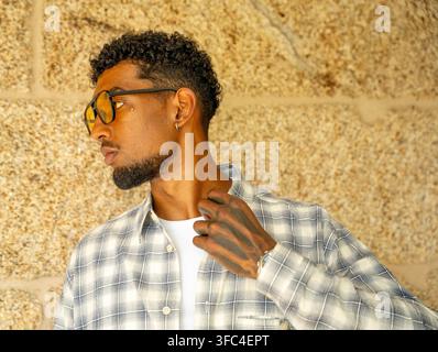 Giovane uomo elegante con occhiali da sole e camicia a quadri che posa di profilo su uno sfondo texturizzato caldo Foto Stock