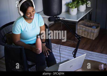 Donna afroamericana con auricolari che tiene una bottiglia rosa, registrando su un laptop con microfono vicino a piante Foto Stock