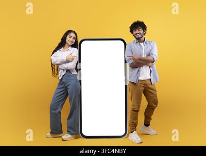 Giovane coppia diversificata e felice che indica uno smartphone con schermo vuoto, sfondo giallo, modello Foto Stock