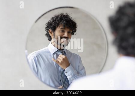 Giovane uomo sorridente che aggiusta la cravatta davanti allo specchio, concetto aziendale Foto Stock