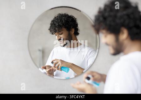 Giovane uomo con capelli ricci che applica un prodotto per la cura davanti allo specchio del bagno, espressione felice Foto Stock
