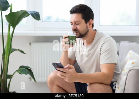 Uomo con smartphone che beve un frullato verde da un bicchiere al chiuso Foto Stock