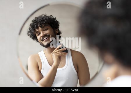 Giovane uomo sorridente che si rade con un rasoio elettrico davanti allo specchio, concetto di cura in bagno Foto Stock