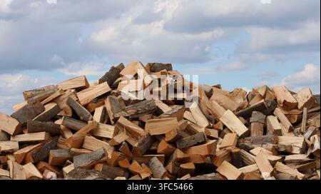 Un grande pila di legna da ardere appena spaccata riempie lo sfondo sotto un cielo parzialmente nuvoloso, mostrando varie texture del legno, corteccia e superfici tagliate in una ruggine Foto Stock
