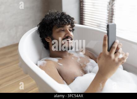 Giovane uomo sudasiatico sorridente che si rilassa nella vasca da bagno mentre usa lo smartphone, concetto di benessere Foto Stock