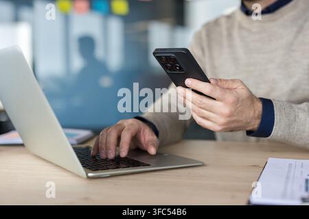 Uomo d'affari che digita contemporaneamente su una tastiera del laptop e tiene uno smartphone, multitasking con la tecnologia per il lavoro da remoto e una comunicazione efficiente in un ambiente d'ufficio Foto Stock