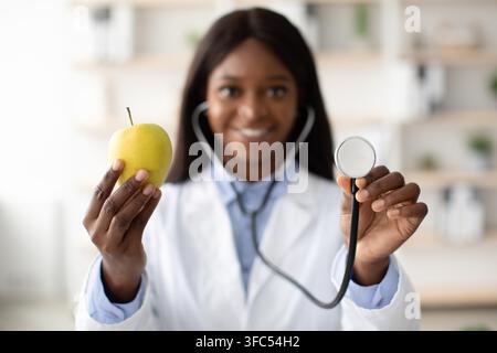 Medica nera felice che tiene una mela e uno stetoscopio, clinica. Concetto di assistenza sanitaria Foto Stock