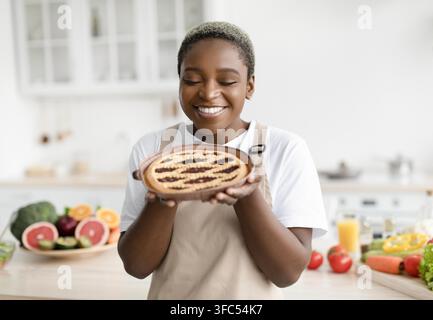 Panificatrice nera sorridente che tiene una torta fatta in casa in una cucina luminosa, esperienza culinaria gioiosa Foto Stock