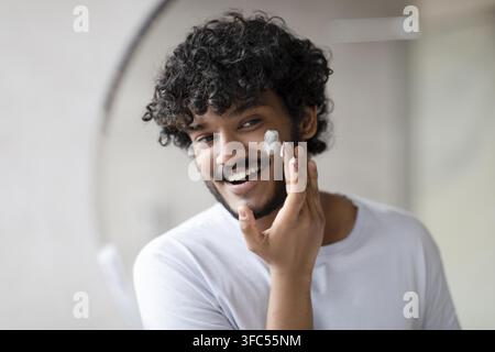 Giovane uomo sorridente con capelli ricci che applica crema per la cura a casa, concetto di bellezza Foto Stock