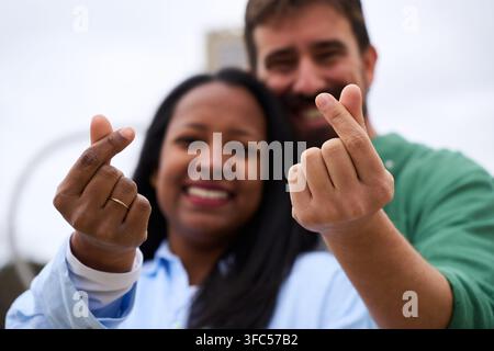 Coppia interrazziale che fa il segno del cuore con le dita in stile coreano mentre guarda dritto nella fotocamera sorridendo. Foto Stock
