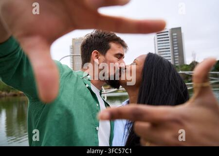 Coppia multirazziale felice che si bacia mentre scatta una foto formando una cornice con le mani. Foto Stock