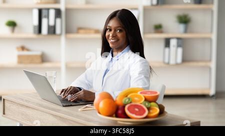 Nutrizionista nera sorridente che lavora su un laptop in ufficio, concetto di assistenza sanitaria Foto Stock