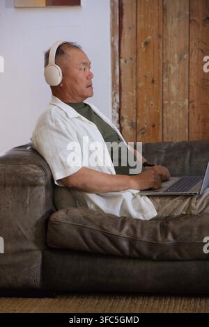 Uomo anziano asiatico seduto su un divano di pelle marrone scuro, con le cuffie e che digita su un laptop Foto Stock