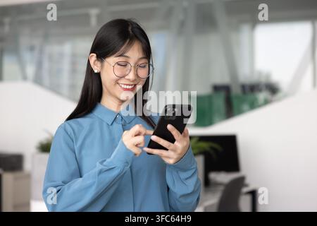 Giovane donna asiatica sorridente con occhiali che utilizza uno smartphone in ufficio Foto Stock