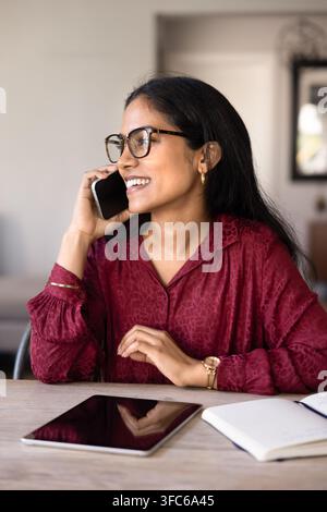 Giovane donna indiana che parla al smartphone, seduta a tavola Foto Stock