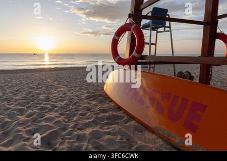 Panneggio di salvataggio del bagnino appoggiato a una torre di legno con anelli di salvataggio su una spiaggia sabbiosa all'alba, luce dorata calda sulle acque calme dell'oceano che creano una pe Foto Stock