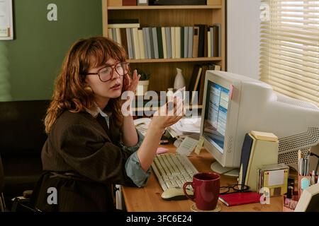 Donna che guarda nello specchio a una scrivania da ufficio retrò con computer vintage Foto Stock