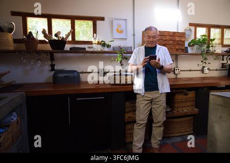 Uomo asiatico anziano in piedi al bancone della cucina, che usa uno smartphone vicino a un piano di lavoro in legno e piante Foto Stock