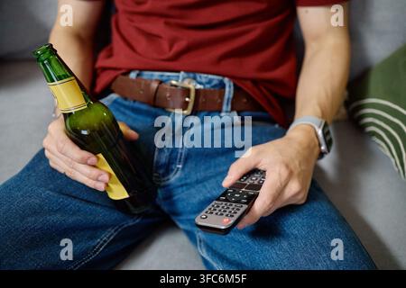 Inquadratura ritagliata di un uomo che tiene una bottiglia di birra e un telecomando TV Foto Stock