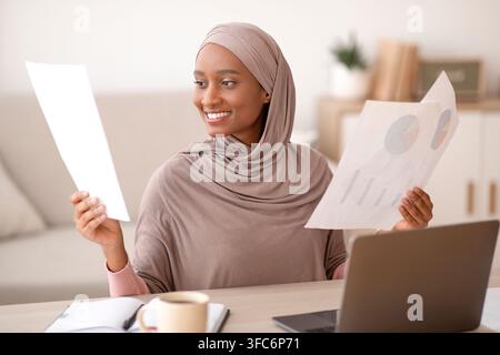 Concetto di lavoro da casa. Felice donna musulmana nera in hijab guardando attraverso i documenti di fronte al computer portatile all'interno Foto Stock