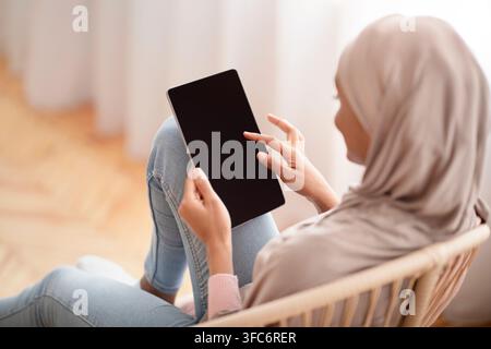 Donna araba nera in hijab che usa un tablet con schermo vuoto mentre si siede in una comoda poltrona a casa, mockup per il design Foto Stock