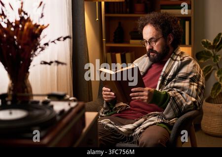 Uomo di mezza età che legge un libro in una comoda poltrona a casa Foto Stock