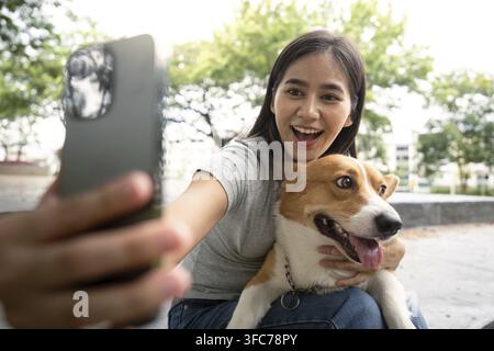 Una giovane donna scatta un selfie gioioso con il suo corgi e celebra la fortuna di possedere animali domestici in un vivace parco Foto Stock
