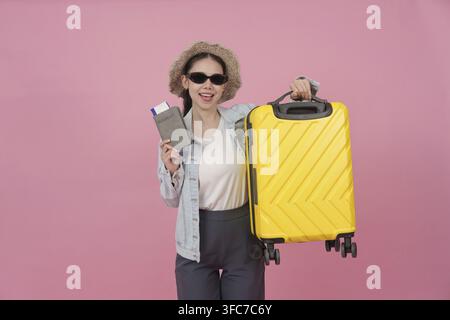 Una viaggiatrice asiatica felice che indossa occhiali da sole e un cappello tiene pronto un passaporto, biglietti aerei e una valigia gialla per il suo viaggio Foto Stock