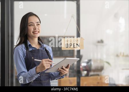 Bellissima giovane barista asiatica in grembiule che tiene un tablet e si trova fuori dalla porta di un bar con un cartello aperto. Concetto di startup per bu Foto Stock