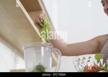In una cucina moderna, una donna mette verdure fresche in un frullatore e prepara un frullato nutriente. Concetto per un'alimentazione sana e il benessere Foto Stock