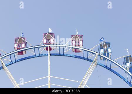 Fragmento di una ruota panoramica contro un cielo azzurro, con cabine rosa e blu e strutture di supporto bianche Foto Stock