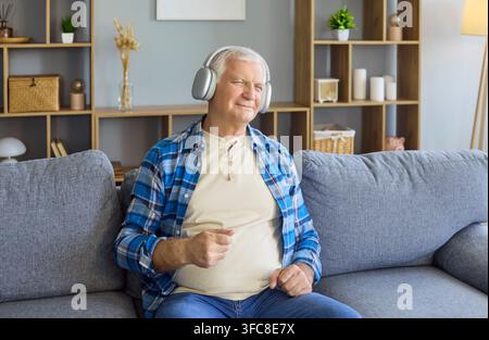 Uomo anziano soddisfatto che ascolta musica a casa sul divano con le cuffie comodamente Foto Stock