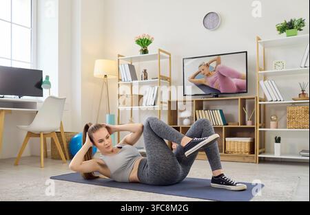 Giovane donna che fa addominali in bicicletta a casa, allenamento online con allenatore TV Foto Stock