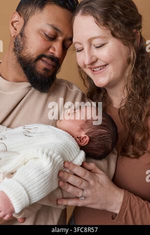 Genitori multirazziali teneri che tengono un neonato in un ritratto familiare caldo, abbracciando la genitorialità e l'amore Foto Stock