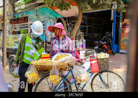 Phnom Penh, Cambogia – 13 febbraio 2020: Vista di una venditrice ambulante che vende frutta dalla sua bicicletta mentre interagisce con un operaio con casco e alto… Foto Stock