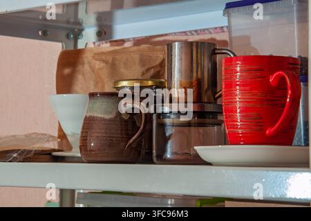 Varie tazze da caffè, un barattolo di fondi di caffè e altri oggetti da cucina sono disposti ordinatamente su una mensola di metallo Foto Stock