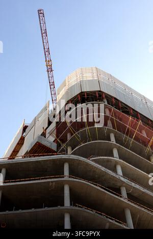 Vista a bassa angolazione di un edificio in cemento a più piani in costruzione, con piani esposti, reti di sicurezza, supporti temporanei e una gru a torre ancora Foto Stock