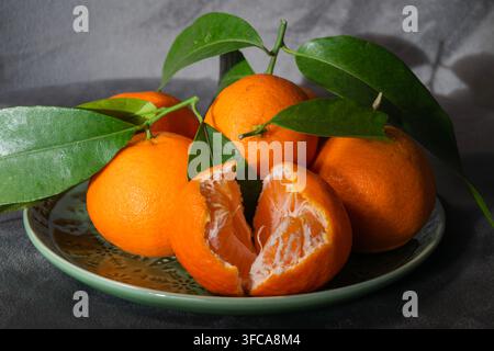 Mandarini freschi e maturi con foglie verdi sono disposti su un piatto decorativo, incluso uno aperto per mostrare i suoi segmenti. Foto Stock