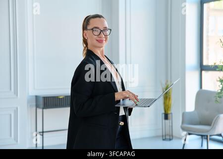 Donna d'affari sorridente con occhiali che tiene un laptop in piedi in un ufficio moderno Foto Stock