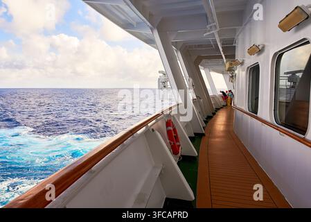 Passeggeri in piedi sulla ringhiera del ponte passeggiata di una nave da crociera che naviga attraverso le acque blu dell'oceano sotto un cielo parzialmente nuvoloso. Foto Stock