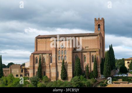 Vista laterale esterna della massiccia basilica di mattoni di San Domenico circondata da cipressi sotto un cielo nuvoloso a Siena, Toscana, Italia. Concetto per Go Foto Stock