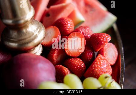 Macro primo piano di fragole rosse mature affettate circondate da fette di anguria e uva su un supporto decorativo dorato per servire Foto Stock