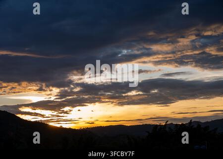 Il sole che tramonta dietro montagne e colline lontane, proiettando una calda luce dorata sul cielo con drammatiche nuvole scure, formando una bella paesaggio naturale Foto Stock