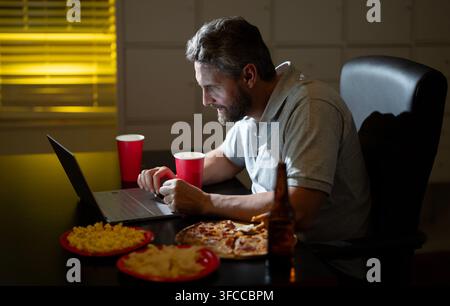 Uomo che mangia popcorn in ufficio di notte. Uomo che mangia snack mentre lavora fino a tardi. Uomo che mangia pizza durante il lavoro notturno. Uomo che mangia cibo spazzatura alla scrivania dell'ufficio Foto Stock