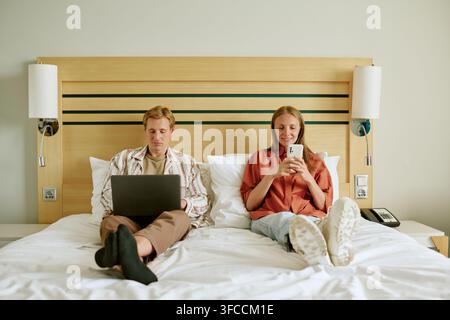 Uomo e donna che usano un laptop e uno smartphone nel letto d'albergo Foto Stock