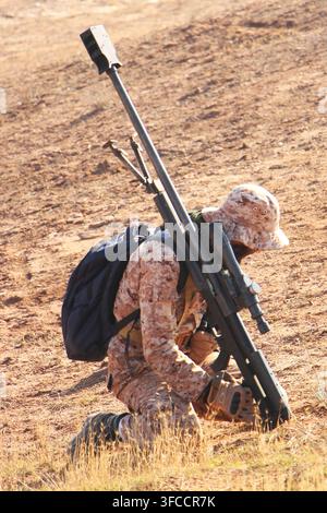 Soldato cecchino militare con mimetica desertica in ginocchio e che trasporta un pesante fucile cecchino anti-materiale in un campo arido Foto Stock