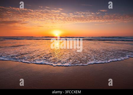 Tramonto dorato sull'oceano sopra una spiaggia sabbiosa con onde gentili e acqua schiumosa Foto Stock