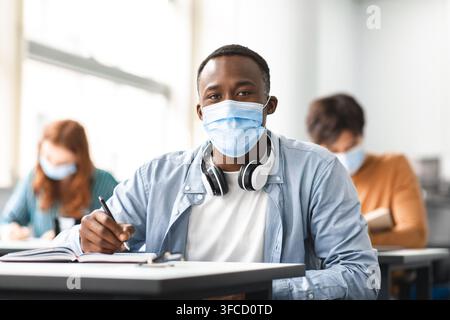 Studente di sesso maschile nero in maschera seduto alla scrivania in classe Foto Stock