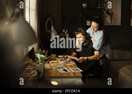 Giovane coppia diversa seduta alla scrivania e che guarda insieme foto stampate. L'immagine rappresenta un hobby creativo e la condivisione di ricordi, adatta al marketing dello stile di vita Foto Stock