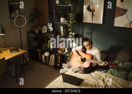 Giovane donna caucasica con cuffie che suona chitarra acustica in camera da letto mentre guarda il laptop. Immagine ideale per lezioni di musica online o marketing di hobby creativi, vista ad angolo alto Foto Stock
